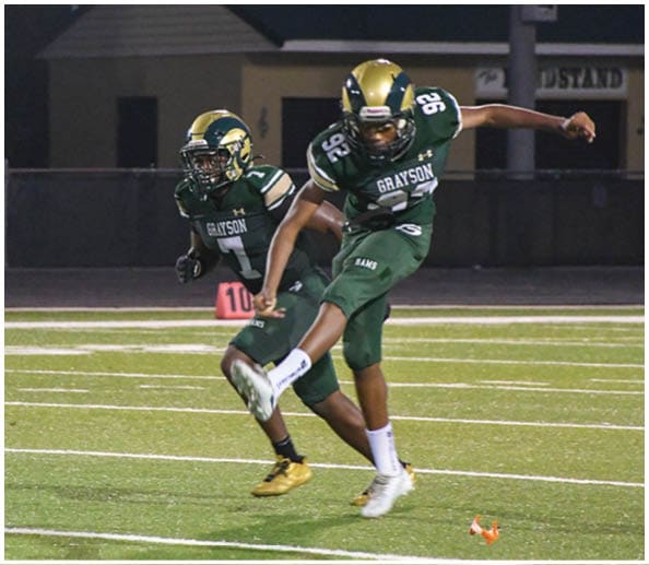 A person in a white and gold football uniform is crouched in an athletic stance on a green football field with other players and a coach in the background. Description generated by AI
