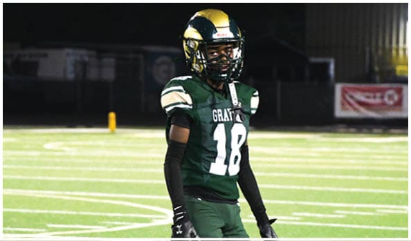 A person in a white and green football uniform with a gold helmet is on a field facing away from the camera while another person in a maroon and gold uniform stands in the background near empty bleachers. Description generated by AI