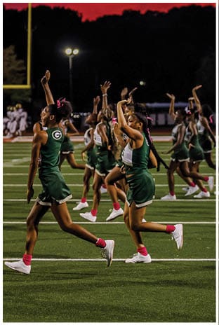 A group of football players in white uniforms and helmets run energetically through a torn banner held by cheerleaders on a field. Description generated by AI