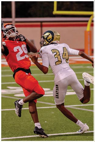 A group of football players in white uniforms and helmets run energetically through a torn banner held by cheerleaders on a field. Description generated by AI
