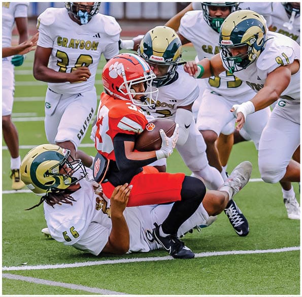 A group of football players in white uniforms and helmets run energetically through a torn banner held by cheerleaders on a field. Description generated by AI