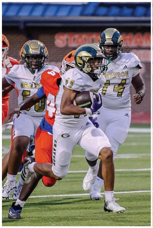 A group of football players in white uniforms and helmets run energetically through a torn banner held by cheerleaders on a field. Description generated by AI