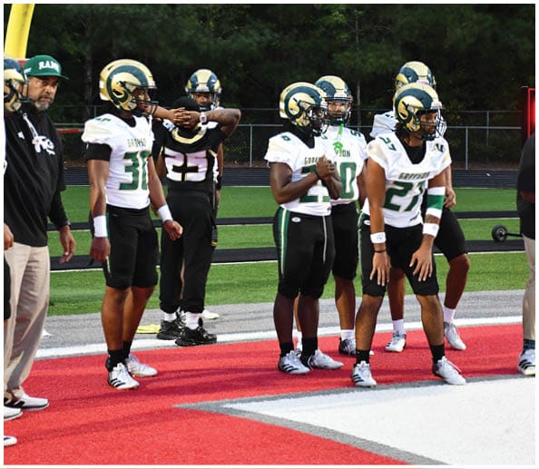 A person in a white and gold football uniform is crouched in an athletic stance on a green football field with other players and a coach in the background. Description generated by AI