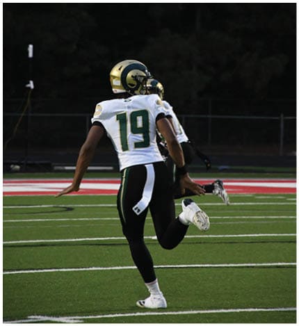 A person with long dreadlocks wearing a white football jersey with the name Grayson and the number 28 stands in front of two other people in similar football uniforms and helmets. Description generated by AI