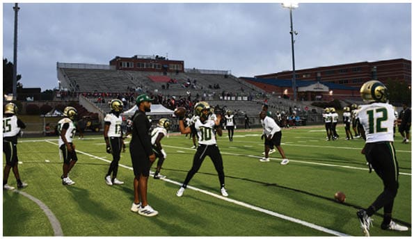 A group of football players in white uniforms and helmets run energetically through a torn banner held by cheerleaders on a field. Description generated by AI