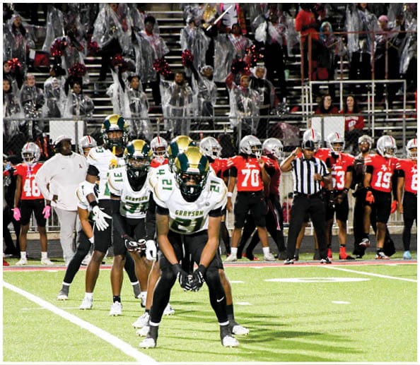 A person in a white and green football uniform with a gold helmet is on a field facing away from the camera while another person in a maroon and gold uniform stands in the background near empty bleachers. Description generated by AI