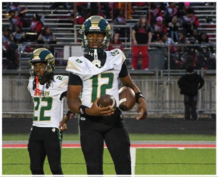 A person wearing a white and gold football uniform with the number 20 stands on a field while two other people in red and gold football uniforms are visible in the background near empty bleachers. Description generated by AI