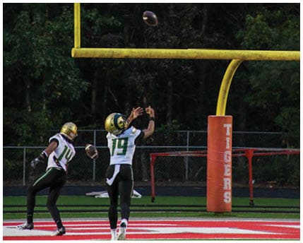 A person wearing a white and yellow football uniform with the number 16 is standing on a field near bleachers where two people are seated. Description generated by AI