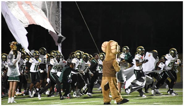 A person wearing a white football uniform and helmet is running with a football on a field while people in green cheerleading outfits stand in the background. Description generated by AI