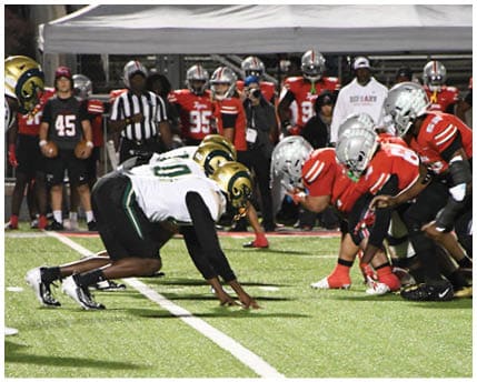 Three people wearing white and gold football uniforms and helmets are standing and interacting near a chain-link fence with trees and bleachers in the background. Description generated by AI