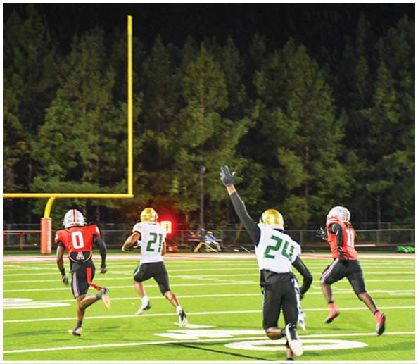 A person in a white and gold football uniform is crouched in an athletic stance on a green football field with other players and a coach in the background. Description generated by AI