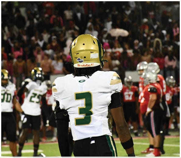A person in a white and green football uniform with a gold helmet is on a field facing away from the camera while another person in a maroon and gold uniform stands in the background near empty bleachers. Description generated by AI
