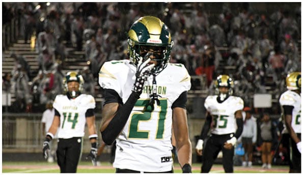 A person wearing a white football uniform and helmet is running with a football on a field while people in green cheerleading outfits stand in the background. Description generated by AI