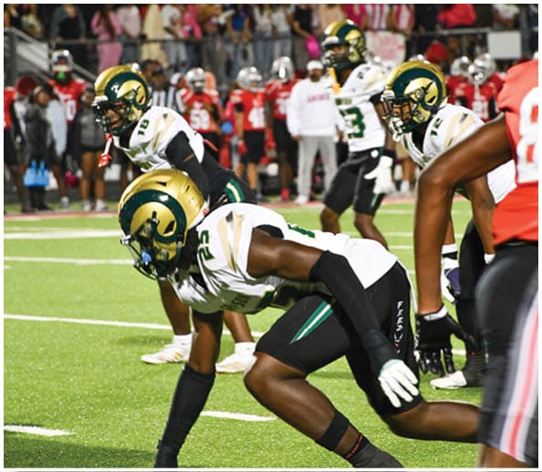 A person in a white and gold football uniform is crouched in an athletic stance on a green football field with other players and a coach in the background. Description generated by AI