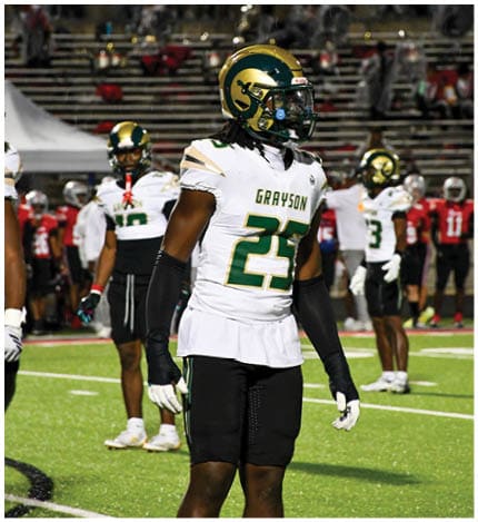 A person with long dreadlocks wearing a white football jersey with the name Grayson and the number 28 stands in front of two other people in similar football uniforms and helmets. Description generated by AI