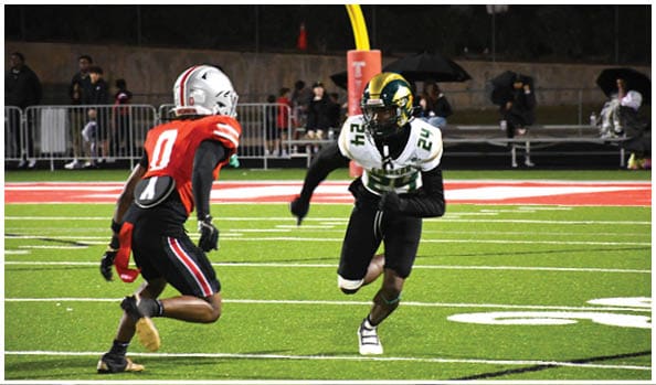 A person in a white and green football uniform with a gold helmet is on a field facing away from the camera while another person in a maroon and gold uniform stands in the background near empty bleachers. Description generated by AI