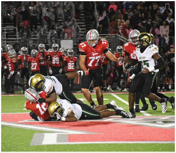 A person in a white and gold football uniform is crouched in an athletic stance on a green football field with other players and a coach in the background. Description generated by AI