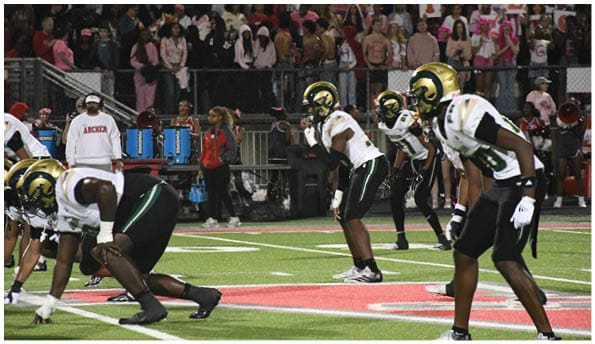 A group of football players in white uniforms and helmets run energetically through a torn banner held by cheerleaders on a field. Description generated by AI