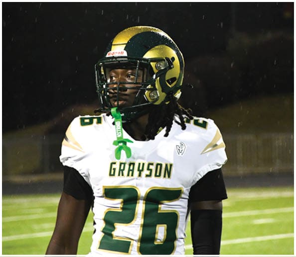 A person in a white and green football uniform with a gold helmet is on a field facing away from the camera while another person in a maroon and gold uniform stands in the background near empty bleachers. Description generated by AI