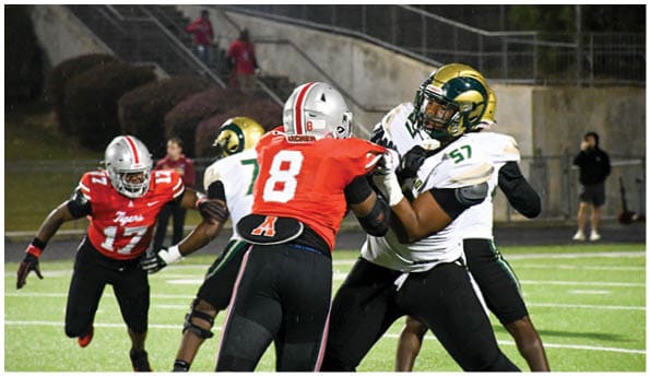 A person wearing a white football uniform and helmet is running with a football on a field while people in green cheerleading outfits stand in the background. Description generated by AI