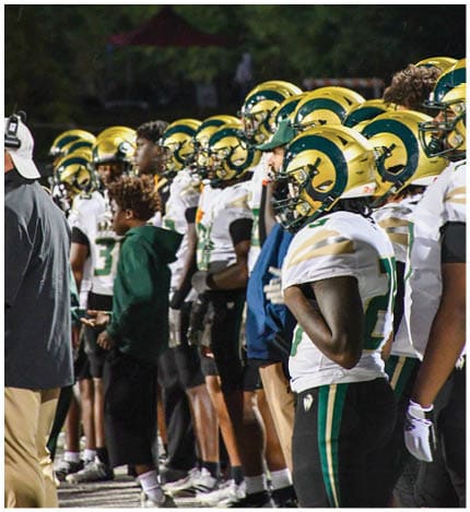 A person with long dreadlocks wearing a white football jersey with the name Grayson and the number 28 stands in front of two other people in similar football uniforms and helmets. Description generated by AI