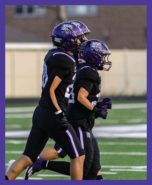 Two people in green football uniforms kneel on a field holding their helmets with heads bowed while cheerleaders stand blurred in the background. Description generated by AI