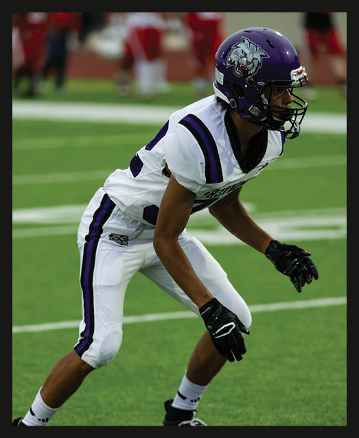 A person with curly hair wearing a black football uniform with “VISTA" on the jersey is sitting on a grassy field with one leg bent and the other extended. Description generated by AI