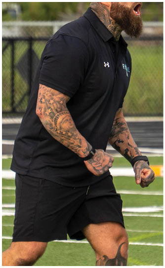 A man with tattoos and a black shirt is standing on a football field. Description generated by AI
