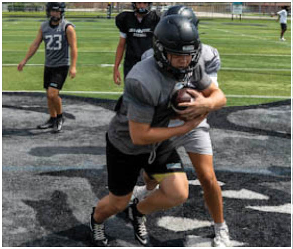 A football player in a grey shirt and black shorts is running with a football. Description generated by AI