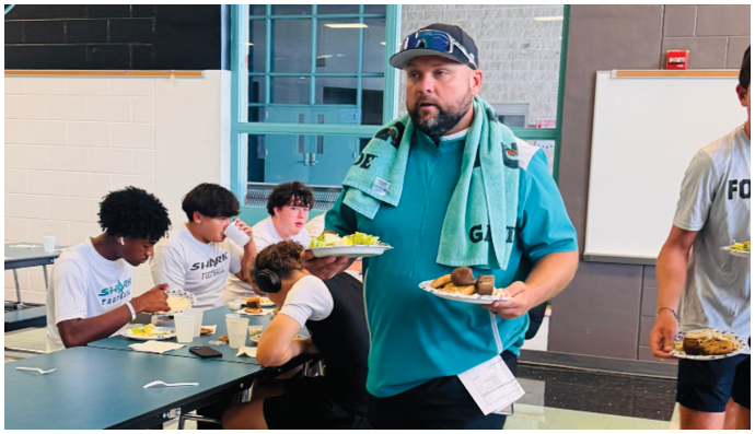 A man wearing a blue shirt and a green scarf is walking through a room with a tray of food. Description generated by AI