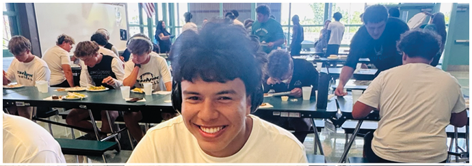 A young man is smiling at the camera while sitting at a table with a plate of food. Description generated by AI