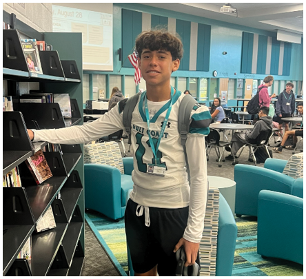A young man wearing a white shirt and black shorts stands in front of a bookshelf. Description generated by AI