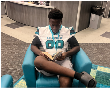 A man is sitting on a blue chair reading a book. Description generated by AI