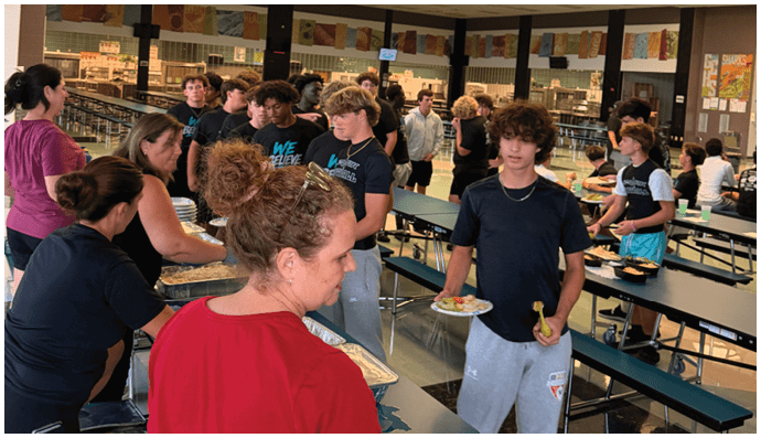 A woman in a red shirt is serving food to people at a table. Description generated by AI