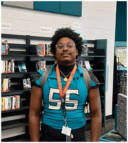 A man wearing a blue jersey with the number 55 standing in front of a bookshelf. Description generated by AI