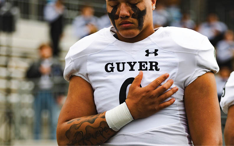 A person in a football uniform with “GUYER" on the jersey stands with eyes closed and a hand placed over their chest. Description generated by AI
