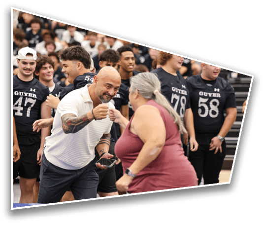 An adult with tattoos and a white shirt is fist bumping another adult with gray hair and a sleeveless top while a group of people in matching sports uniforms watch and smile in the background. Description generated by AI