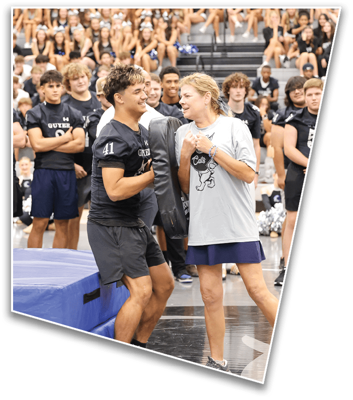 People in athletic clothing are gathered indoors around padded gym mats, with two adults in the foreground appearing to engage in conversation or instruction. Description generated by AI