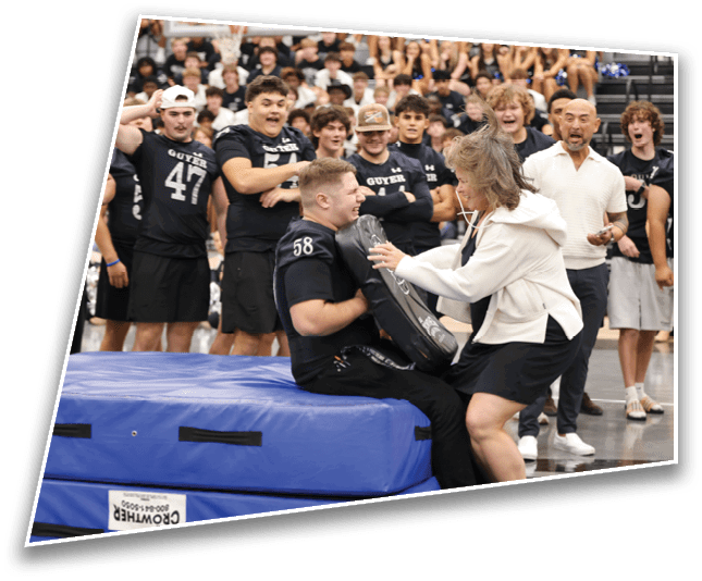 An adult wearing a sports uniform sits on a blue padded mat holding a black padded shield while another adult in a white jacket and black skirt uses a stethoscope on the shield, with a group of people in matching sports uniforms watching in the background. Description generated by AI
