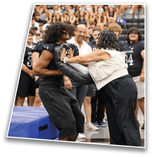 Two people are engaged in a physical training exercise where one person is pushing against a padded blocking sled while others watch in a gymnasium setting. Description generated by AI