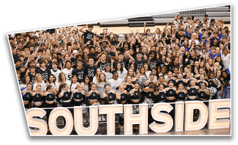 A large group of people wearing matching sports and cheer uniforms are gathered closely together in a gymnasium with bleachers in the background, many smiling and raising their hands. Description generated by AI