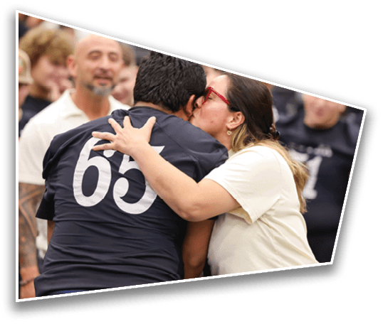 A person wearing a dark shirt with the number 65 is being hugged and kissed on the cheek by another person wearing glasses and a light-colored shirt in a crowded indoor setting. Description generated by AI