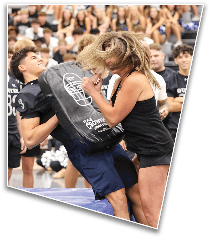 A person with long hair wearing a black tank top and shorts is striking a padded training shield held by another person in a gymnasium with spectators in the background. Description generated by AI