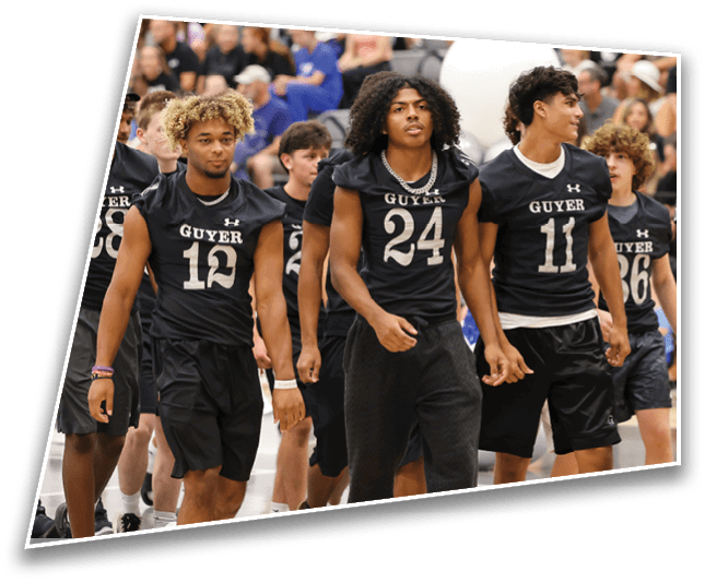 A group of people wearing matching sports jerseys with the word “GUYER" walk together indoors with a crowd seated in the background. Description generated by AI