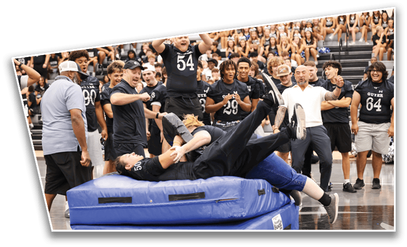 People in sportswear are gathered around two stacked blue mats while one person is falling backward onto the mats during an indoor activity. Description generated by AI