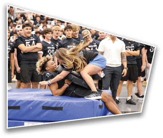 A person in athletic clothing playfully lies on a padded mat while another person in shorts and a t-shirt leans over them, surrounded by a group of people wearing matching sports jerseys in an indoor gym setting. Description generated by AI