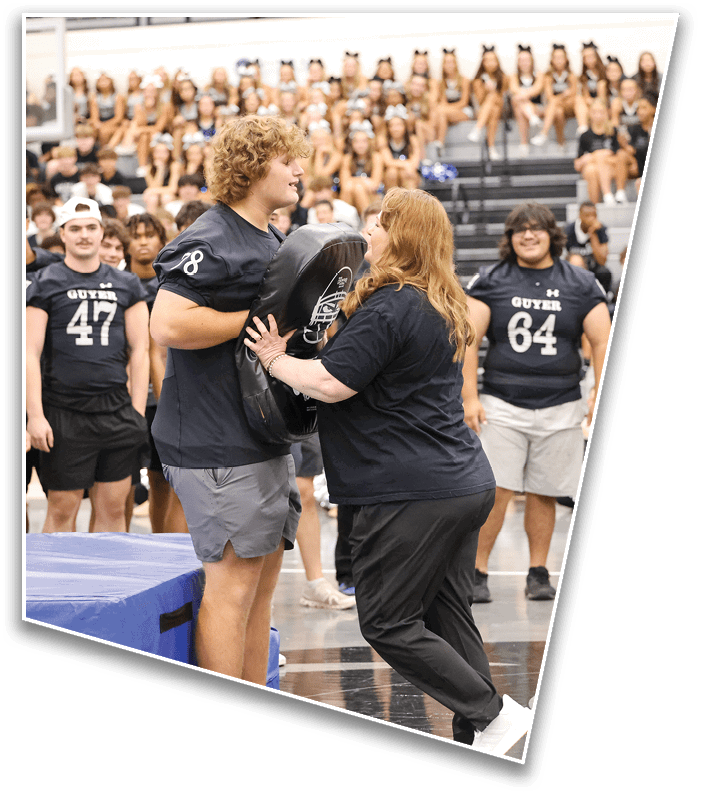 A person with curly hair wearing a sports jersey holds a blocking pad while another person in black athletic clothing pushes against it in a gymnasium with people watching in the background. Description generated by AI