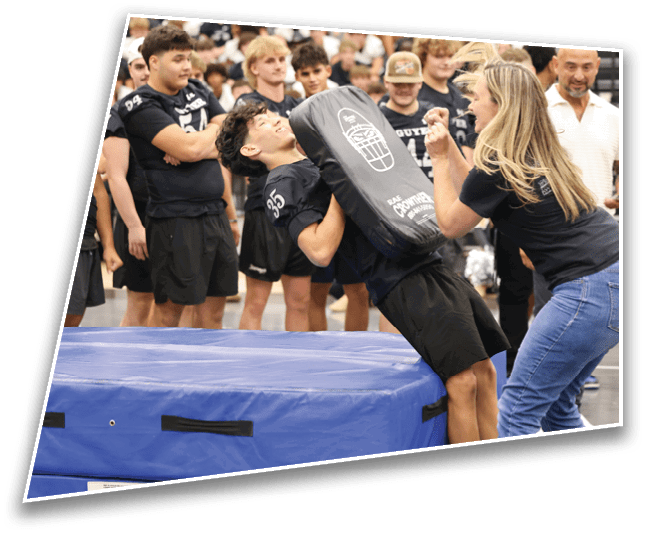 A group of people in sports attire watch as one person leans back against a padded mat while holding a blocking pad and another person gestures energetically in front of them. Description generated by AI