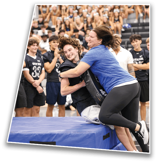 A person in a blue shirt and black leggings is playfully tackling another person with curly hair who is sitting on a padded platform while a group of people in sports uniforms watch and smile in the background. Description generated by AI