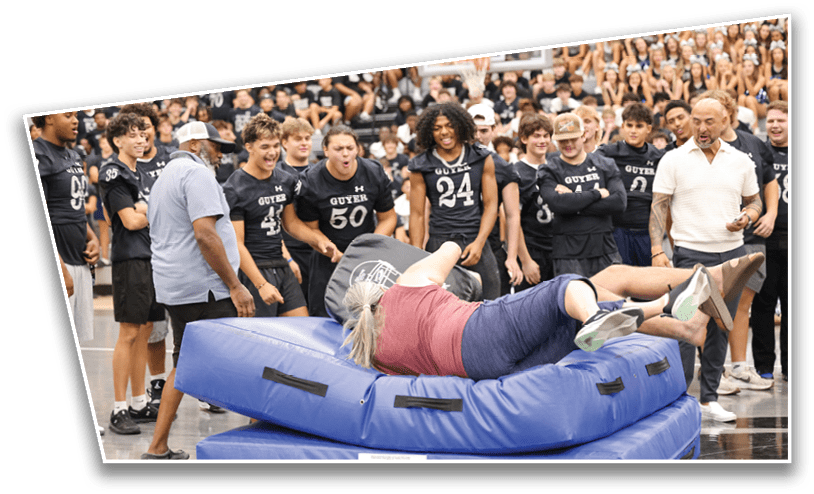 A group of people wearing matching sports jerseys watch and cheer as one person falls backward onto stacked blue gym mats in an indoor gymnasium. Description generated by AI